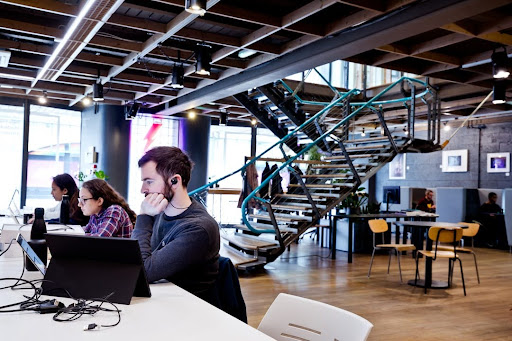 One man and two women seated at their desks looking at computers in a modern, industrial-style office with a large staircase in the background